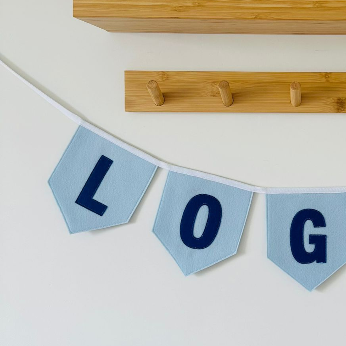 Gepersonaliseerde handgemaakte vilten vlaggenlijn met de letters LOG bestaande uit licht blauw kleurige vlaggen met donker blauw vilten letters. De randen van de vlaggen zijn afgewerkt met een rechte rand. De vlaggenlijn hangt aan een wit katoenen lint.
