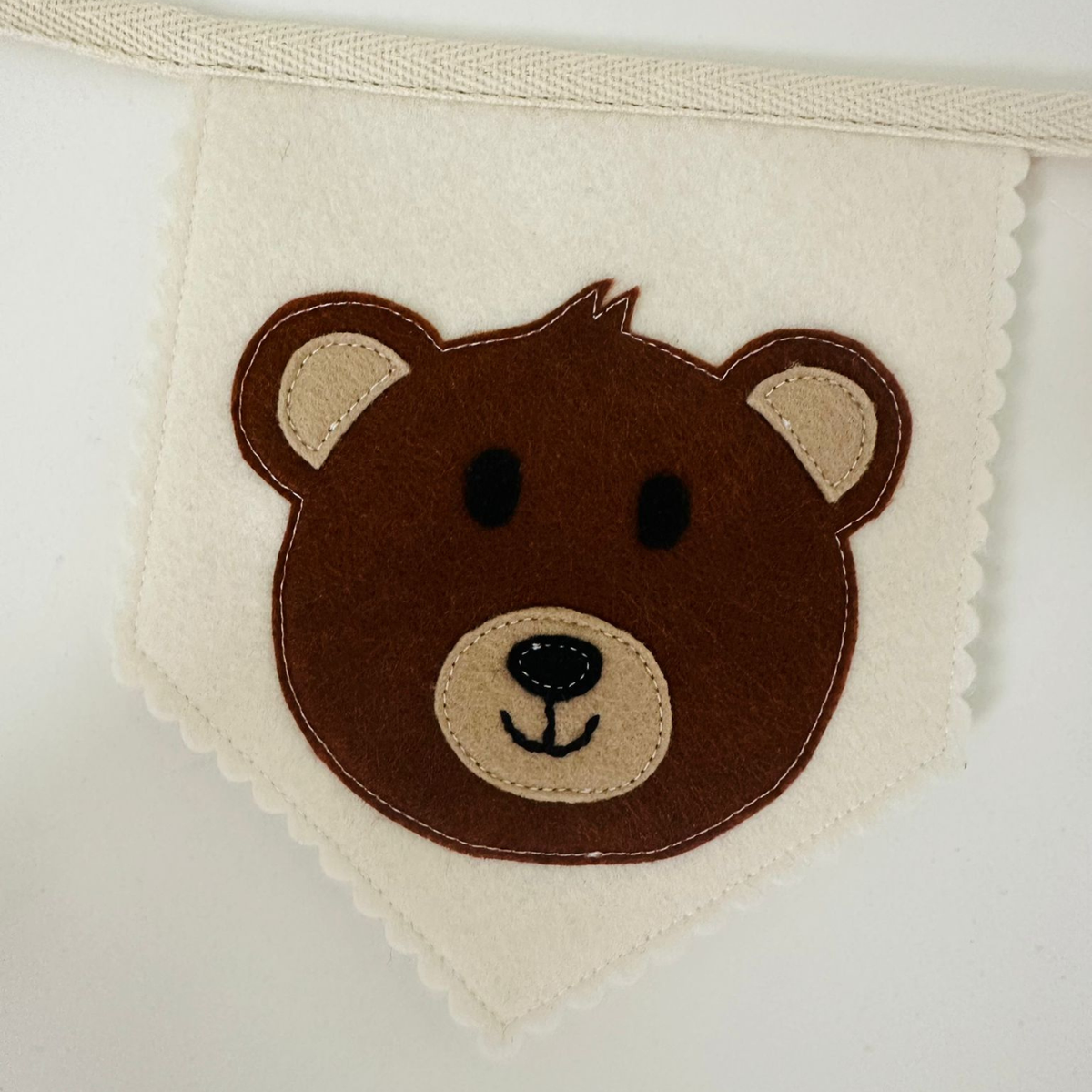 Handgemaakte vilten vlag met een bruine teddybeer icoon op een champagne kleurige vlag, hangend aan een naturel katoenen lint. De beer heeft beige oren en snuit, zwarte ogen, neus en mond. de randen van de vlag zijn met een scallop afwerking afgewerkt.