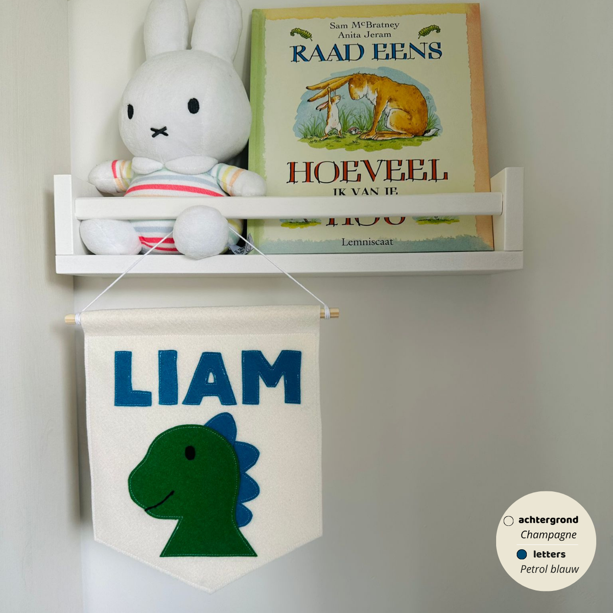 Gepersonaliseerde handgemaakte vilten banner met de naam "Liam", bestaande uit champagne kleurige banner met petroleum blauwe vilten letters en dino hoofd  icoon. De randen van de banner is afgewerkt met een rechte rand. De banner hangt aan een wit katoenen koord aan een houten stok in een kinderkamer.