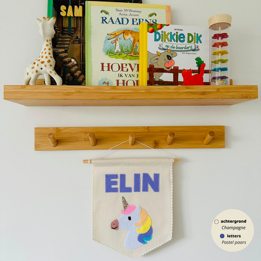 Gepersonaliseerde handgemaakte vilten banner met de naam "Elin", bestaande uit champagne kleurige banner met pastel paars vilten letters en eenhoorn icoon. De randen van de banner is afgewerkt met een scallop rand. De banner hangt aan een wit katoenen koord aan een houten stok in een kinderkamer.
