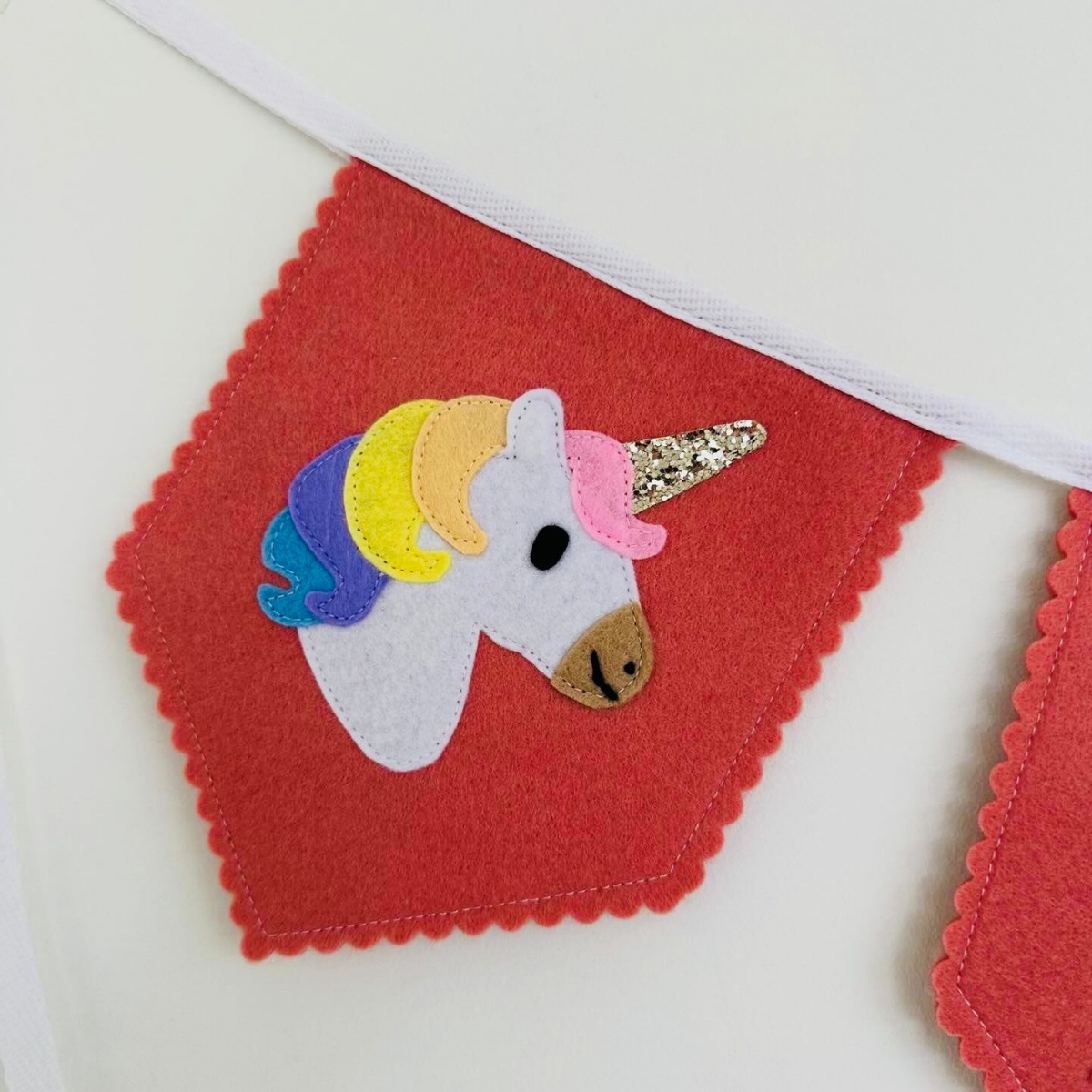 Handgemaakte vilten vlag met een eenhoorn icoon erop, hangend aan een witte katoenen lint. De randen van de vlag zijn met een scalp afwerking afgewerkt.