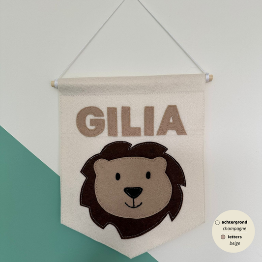 Gepersonaliseerde handgemaakte vilten banner met de naam "GILIA", bestaande uit champagne kleurige banner met beige vilten letters en leeuwenhoofd icoon. De randen van de banner is afgewerkt met een rechte rand. De banner hangt aan een wit katoenen koord aan een houten stok in een kinderkamer.