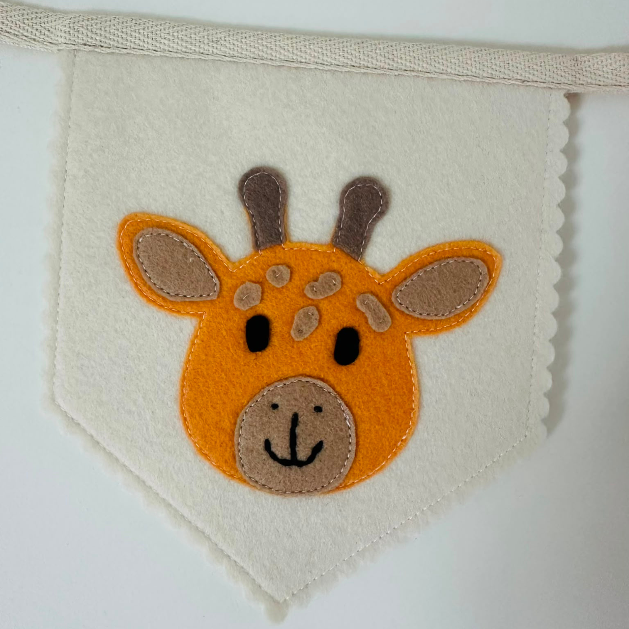 Handgemaakte vilten vlag met een geel giraffenhoofd icoon op een champagne kleurige vlag, hangend aan een naturel katoenen lint. De giraf heeft taupe oren en beige snuit en vlekken, zwarte ogen, neus en mond. de randen van de vlag zijn met een scallop afwerking afgewerkt.