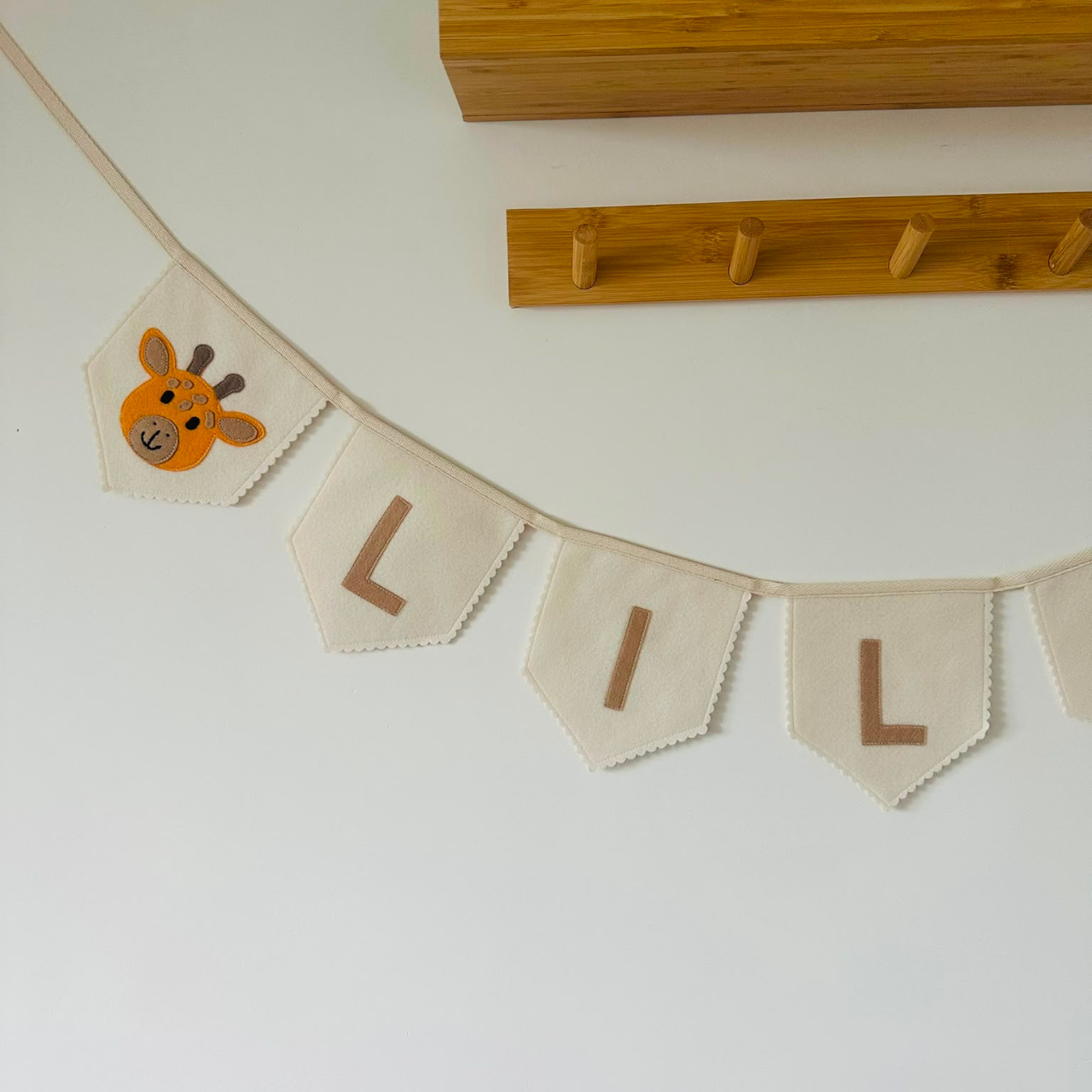 Gepersonaliseerde handgemaakte vilten naamvlaggenlijn met de eerste 3 letters van Milou, bestaande uit champagne kleurige vlaggen met beige vilten letters en een geel giraffenhoofd icoon op de eerste vlag. De randen van de vlaggen zijn afgewerkt met een scallop rand. De vlaggenlijn hangt aan een naturel katoenen lint, gefotografeerd tegen een lichte muur met een houten kapstok op de achtergrond.
