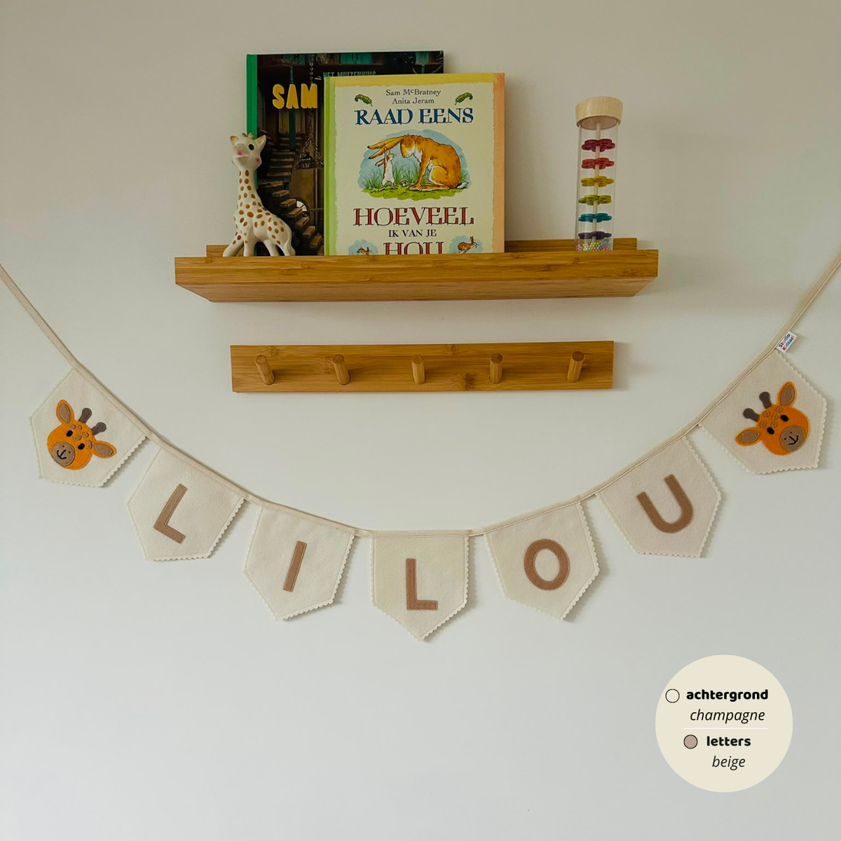 Gepersonaliseerde handgemaakte vilten naamvlaggenlijn met de naam "LILOU", bestaande uit champagne kleurige vlaggen met beige vilten letters en giraffenhoofd iconen op de eerste en laatste vlag. De randen van de vlaggen zijn afgewerkt met een scallop rand. De vlaggenlijn hangt aan een naturel katoenen lint in een kinderkamer, gefotografeerd onder een houten boekenplank met kinderboeken en een Sophie de Giraf speeltje, boven een houten kapstok.
