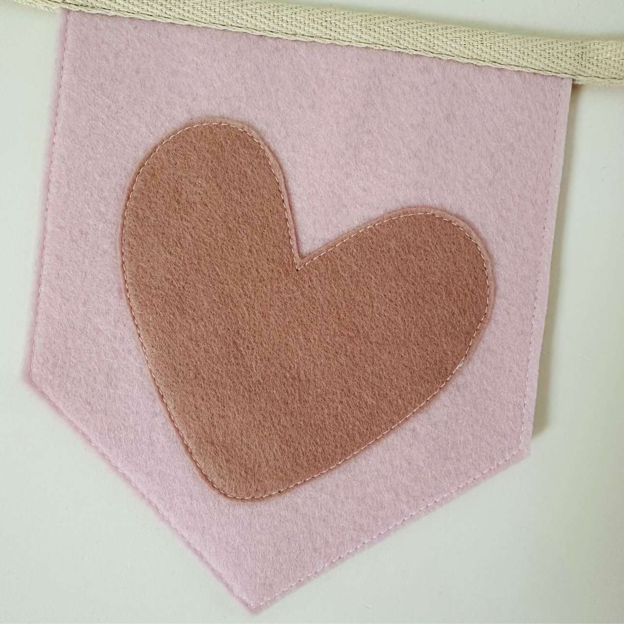 Handgemaakte vilten vlag met een oud roze hart icoon op een licht roze kleurige vlag, hangend aan een naturel katoenen lint. De randen van de vlag zijn met een rechte afwerking afgewerkt.