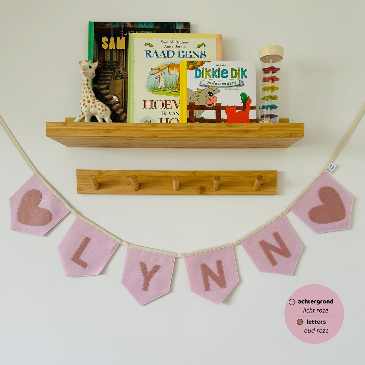 Gepersonaliseerde handgemaakte vilten naamvlaggenlijn met de naam "LYNN", bestaande uit licht roze kleurige vlaggen met oud roze vilten letters en oud roze hart iconen op de eerste en laatste vlag. De randen van de vlaggen zijn afgewerkt met een rechte rand. De vlaggenlijn hangt aan een naturel katoenen lint in een kinderkamer, gefotografeerd onder een houten boekenplank met kinderboeken en een Sophie de Giraf speeltje, boven een houten kapstok.