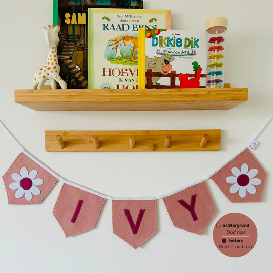 Gepersonaliseerde handgemaakte vilten naamvlaggenlijn met de naam "Ivy", bestaande uit oud roze kleurige vlaggen met donker oud roze vilten letters en wit met donker oud roze bloem op het eerste vlaggetje en het laatste vlaggetje. De randen van de vlaggen zijn afgewerkt met een scalp afwerking. De vlaggenlijn hangt aan een wit katoenen lint.