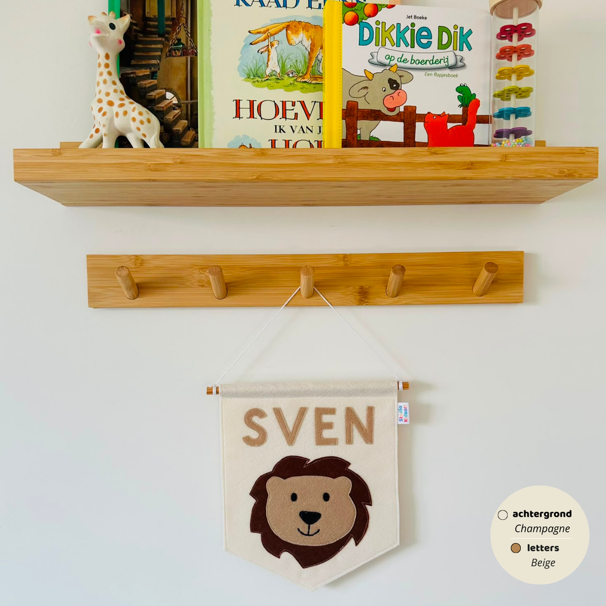 Gepersonaliseerde handgemaakte vilten banner met de naam "Sven", bestaande uit champagne kleurige banner met beige vilten letters en leeuwenhoofd icoon. De randen van de banner is afgewerkt met een rechte rand. De banner hangt aan een wit katoenen koord aan een houten stok in een kinderkamer.