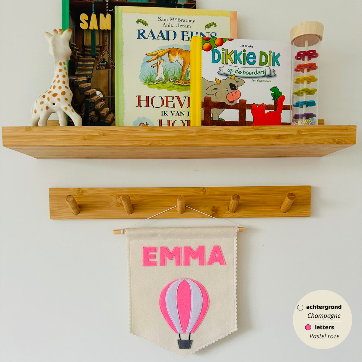 Gepersonaliseerde handgemaakte vilten banner met de naam "Emma", bestaande uit champagne kleurige banner met pastel roze vilten letters en luchtballon icoon. De randen van de banner is afgewerkt met een scallop rand. De banner hangt aan een wit katoenen koord aan een houten stok in een kinderkamer.