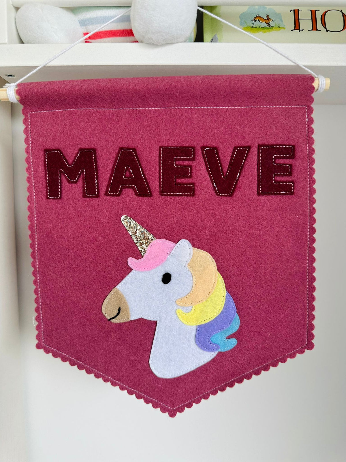 Gepersonaliseerde handgemaakte vilten banner met de naam "Maeve", bestaande uit donker oud roze kleurige banner met bordeaux vilten letters en eenhoorn icoon. De randen van de banner is afgewerkt met een scallop rand. De banner hangt aan een wit katoenen koord aan een houten stok in een kinderkamer.