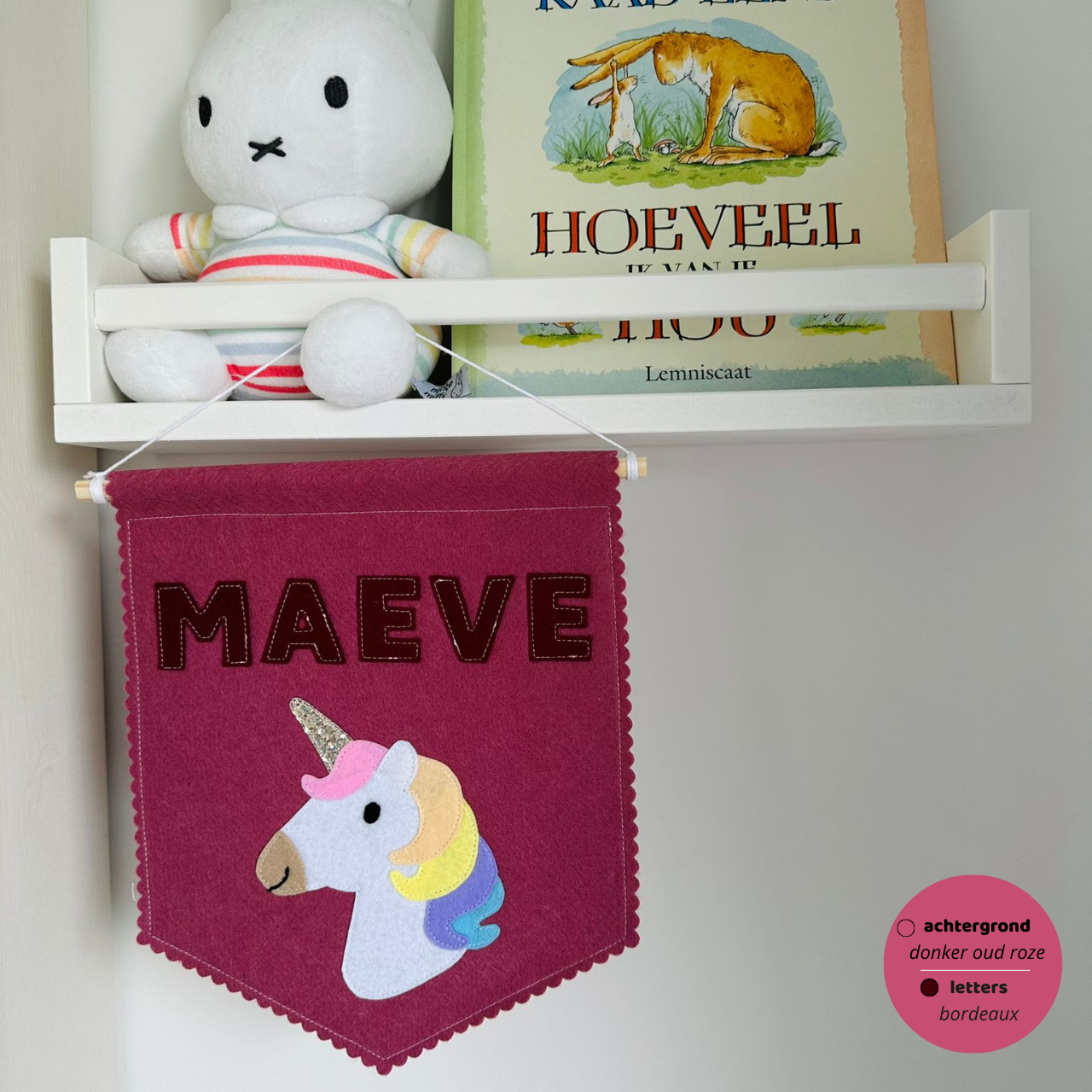 Gepersonaliseerde handgemaakte vilten banner met de naam "Maeve", bestaande uit donker oud roze kleurige banner met bordeaux vilten letters en eenhoorn icoon. De randen van de banner is afgewerkt met een scallop rand. De banner hangt aan een wit katoenen koord aan een houten stok in een kinderkamer.