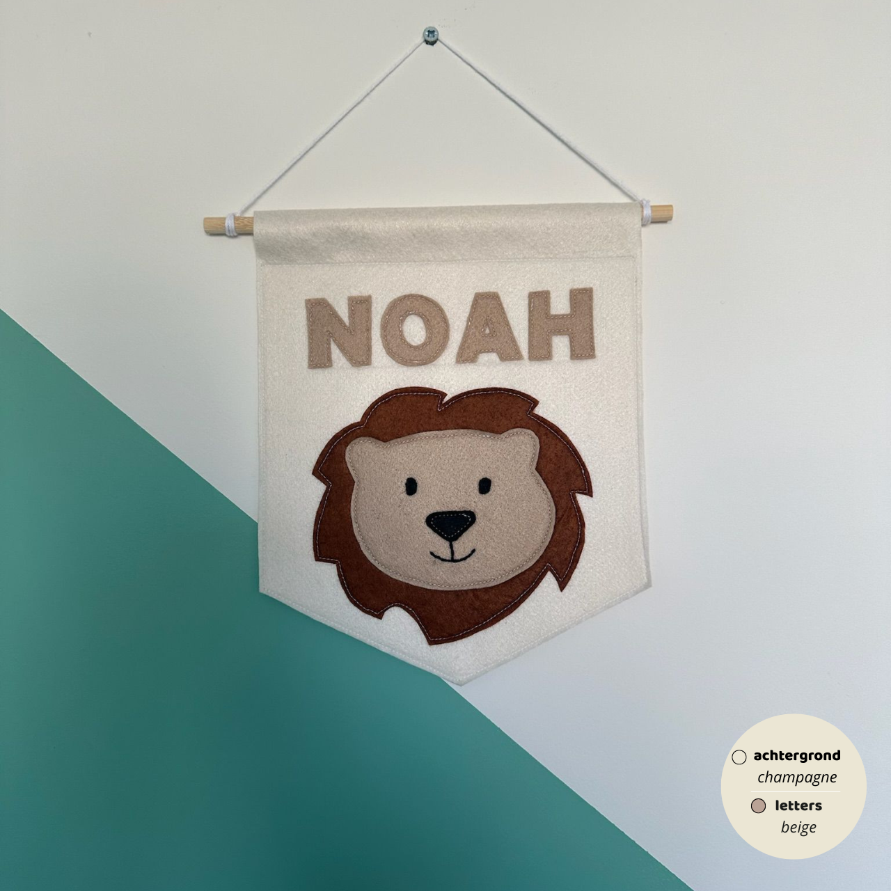 Gepersonaliseerde handgemaakte vilten banner met de naam "NOAH", bestaande uit champagne kleurige banner met beige vilten letters en leeuwenhoofd icoon. De randen van de banner is afgewerkt met een rechte rand. De banner hangt aan een wit katoenen koord aan een houten stok in een kinderkamer.