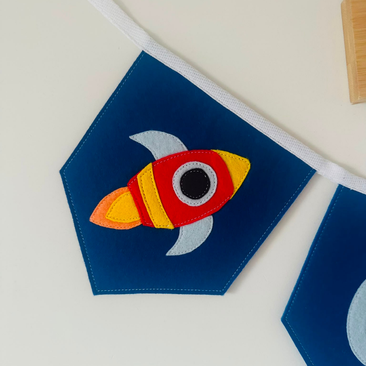 Handgemaakte vilten vlag met een raket icoon erop, hangend aan een wit katoenen lint. De randen van de vlag zijn met een rechte afwerking afgewerkt.