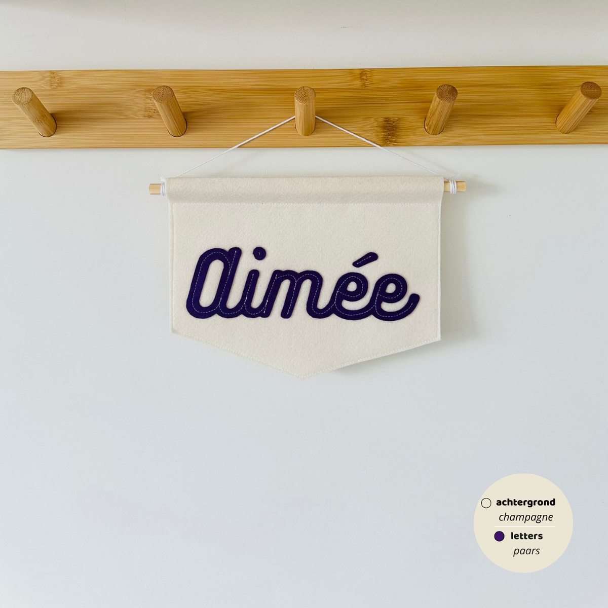 Gepersonaliseerde script banner met de naam 'Aimée'. De letters zijn in lettertype script op de banner. De kleur van de letters zijn paars met een champagne kleurige achtergrond van de banner. De banner hangt aan een houten stokje met wit koord.