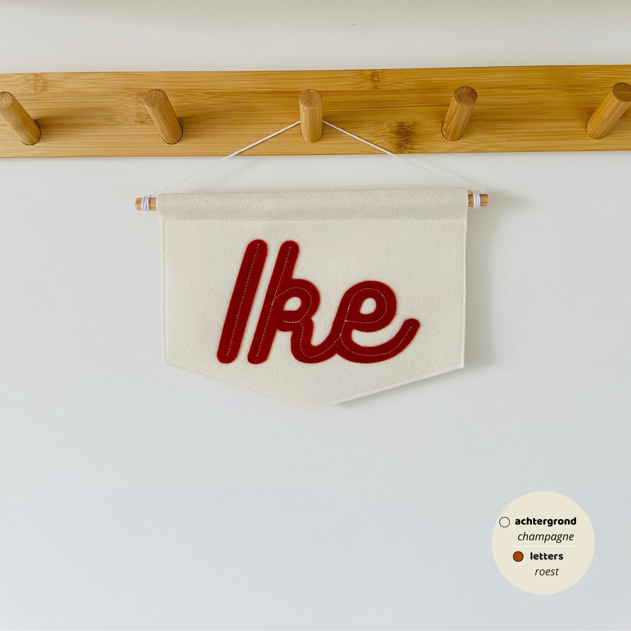 Gepersonaliseerde script banner met de naam 'Ike'. De letters zijn in lettertype script op de banner. De kleur van de letters zijn roest met een champagne kleurige achtergrond van de banner. De banner hangt aan een houten stokje met wit koord.