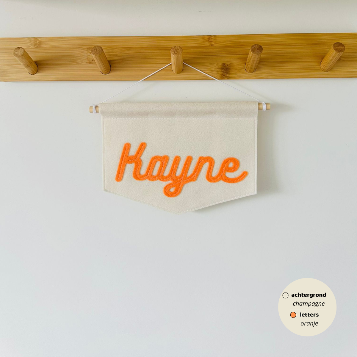 Gepersonaliseerde script banner met de naam 'Kayne'. De letters zijn in lettertype script op de banner. De kleur van de letters zijn oranje met een champagne kleurige achtergrond van de banner. De banner hangt aan een houten stokje met wit koord.