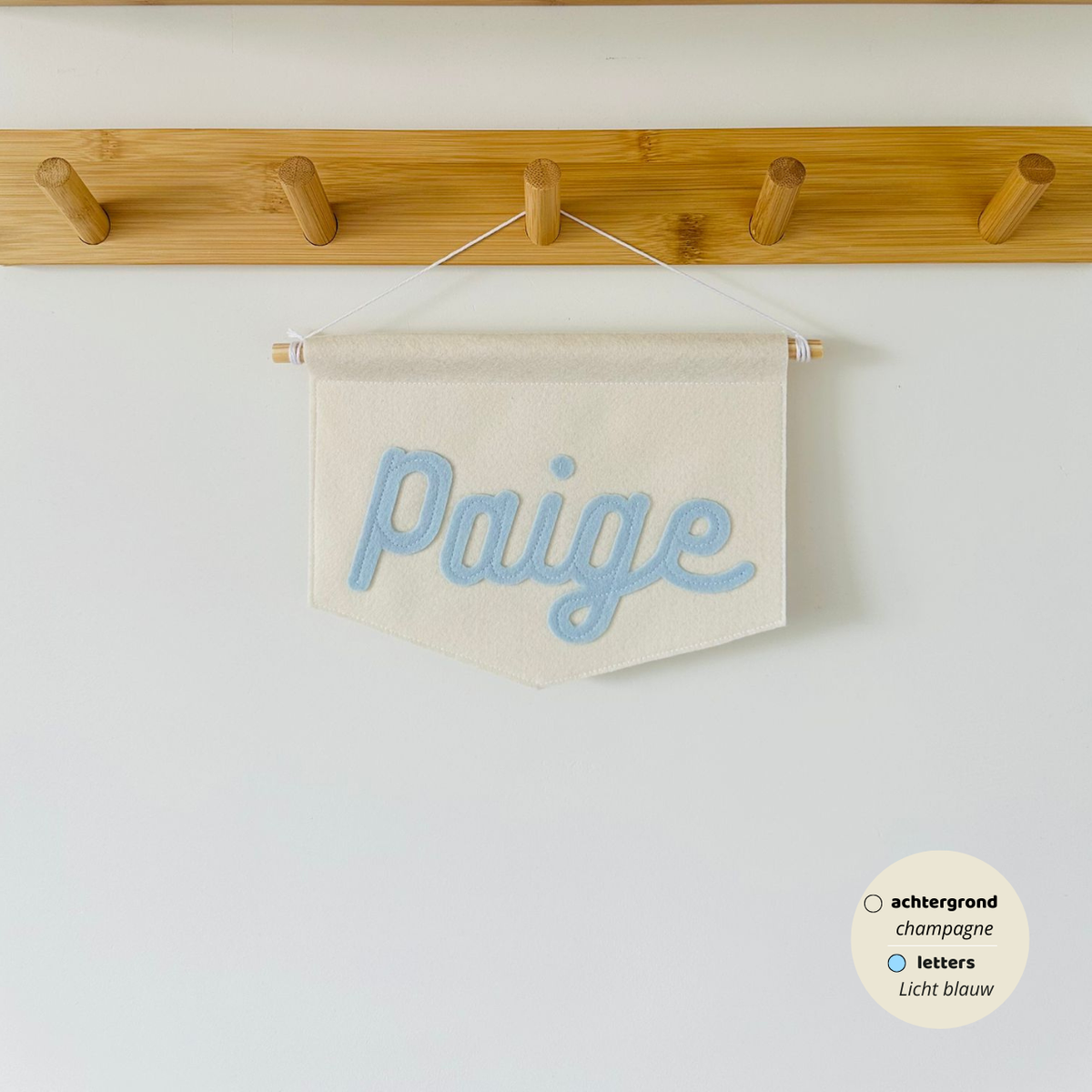 Gepersonaliseerde script banner met de naam 'Paige'. De letters zijn in lettertype script op de banner. De kleur van de letters zijn licht blauw met een champagne kleurige achtergrond van de banner. De banner hangt aan een houten stokje met wit koord.