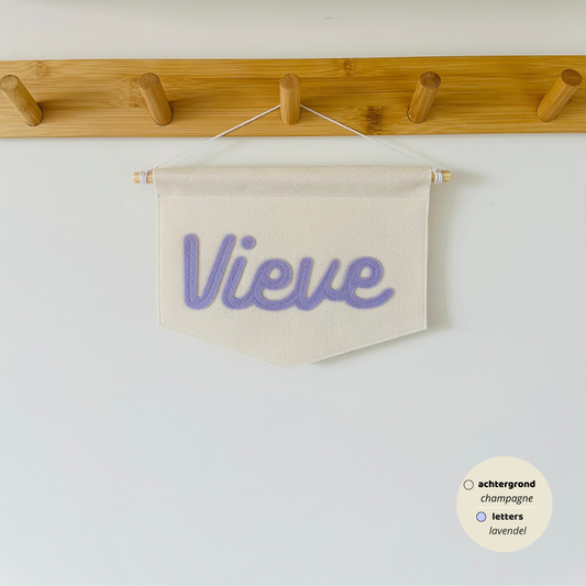 Gepersonaliseerde script banner met de naam 'Vieve'. De letters zijn in lettertype script op de banner. De kleur van de letters zijn lavendel met een champagne kleurige achtergrond van de banner. De banner hangt aan een houten stokje met wit koord.