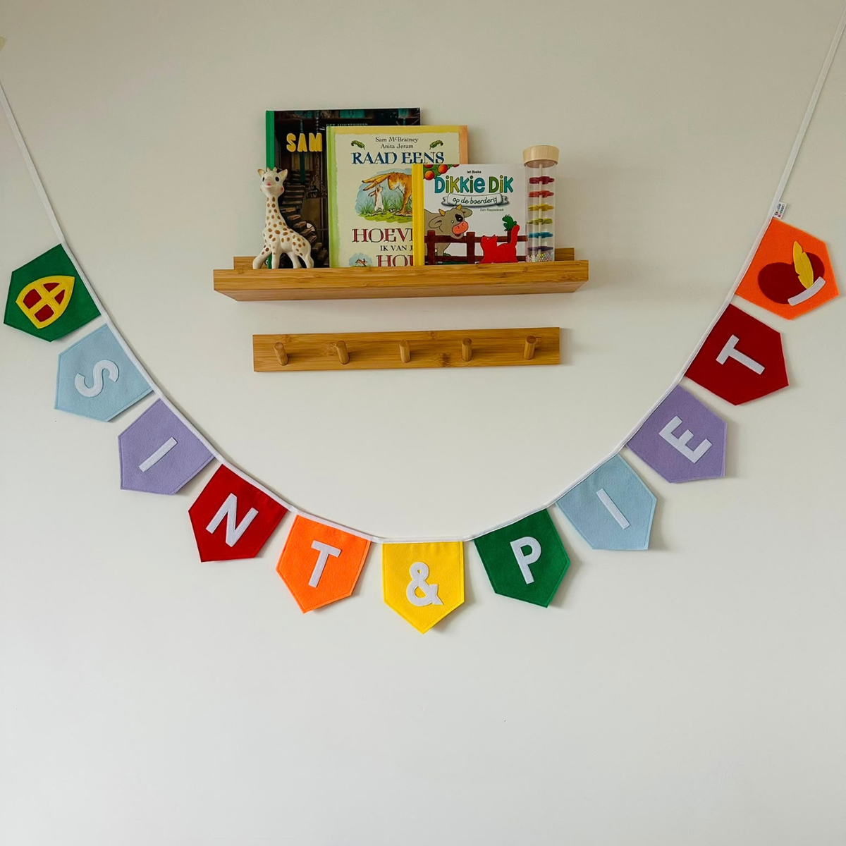 Sinterklaas vlaggenlijn van vilt met het woord SINT & Piet in witte letters op gekleurde vlaggetjes, met aan de uiteinden een mijter en een Pietenmuts, hangend aan een muur boven een houten wandplank