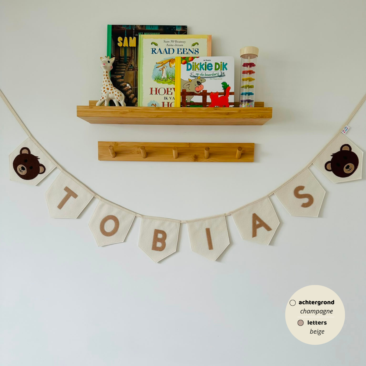 Gepersonaliseerde handgemaakte vilten naamvlaggenlijn met de naam "TOBIAS", bestaande uit champagne kleurige vlaggen met beige vilten letters en bruine teddybeer iconen op de eerste en laatste vlag. De randen van de vlaggen zijn afgewerkt met een rechte rand. De vlaggenlijn hangt aan een naturel katoenen lint in een kinderkamer, gefotografeerd onder een houten boekenplank met kinderboeken en een Sophie de Giraf speeltje, boven een houten kapstok.