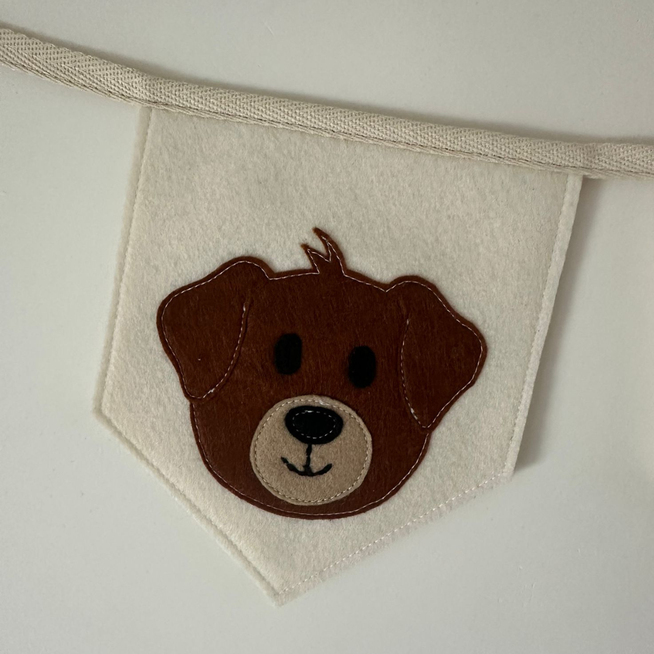 Handgemaakte vilten vlag met bruin hondenhoofd icoonmet beige snoet op een champagne kleurige vlag, hangend aan een naturel katoenen lint. De randen van de vlag zijn met een rechte afwerking afgewerkt.