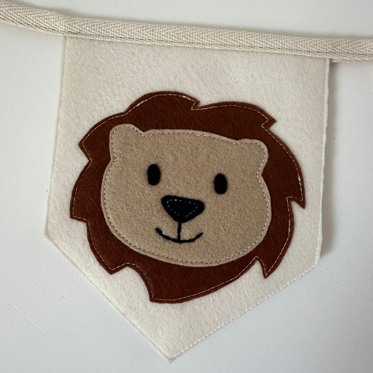 Handgemaakte vilten vlag met een bruin beige leeuwenhoofd icoon op een champagne kleurige vlag, hangend aan een naturel katoenen lint. De leeuw heeft beige gezicht, zwarte ogen, neus en mond. de randen van de vlag zijn met een rechte afwerking afgewerkt.