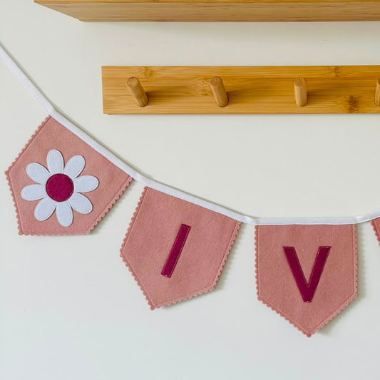 Close-up van een handgemaakt vilten vlaggetje uit een gepersonaliseerde bloem vlaggenlijn, met een oud roze vlaggetje en een witte vilten bloem met donker oud roze hardafgewerkt met een scalloped rand en wit katoenen lint.