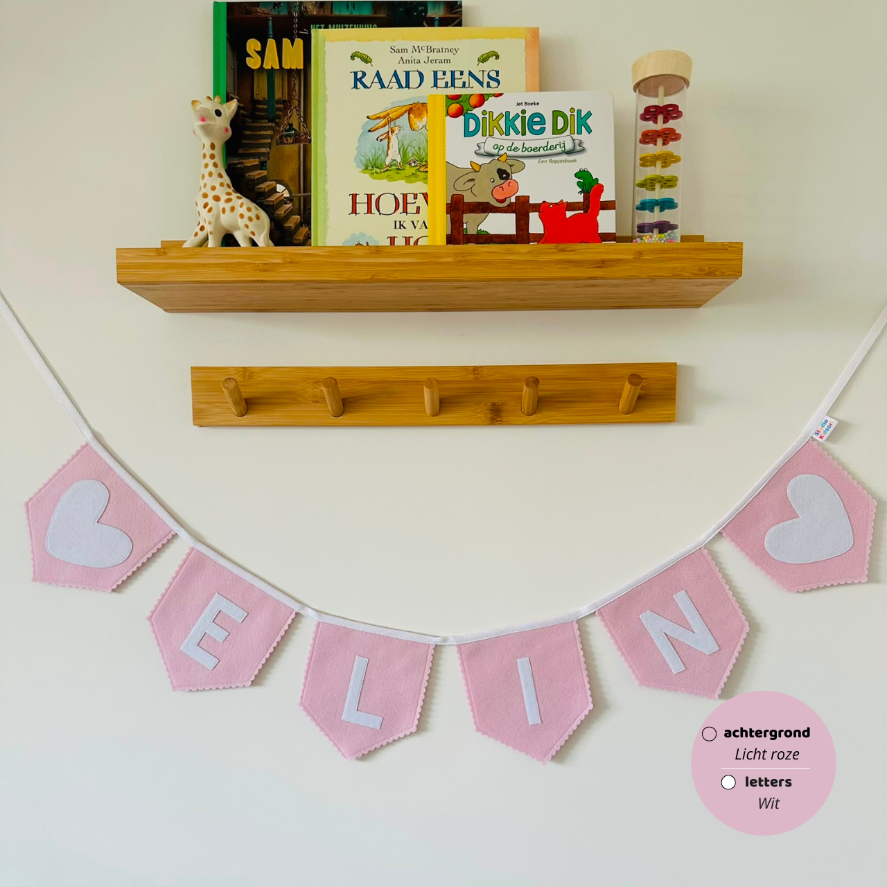 Gepersonaliseerde handgemaakte vilten naamvlaggenlijn met de naam "ELIN", bestaande uit licht roze kleurige vlaggen met wit vilten letters en witte hart iconen op de eerste en laatste vlag. De randen van de vlaggen zijn afgewerkt met een scallop rand. De vlaggenlijn hangt aan een wit katoenen lint in een kinderkamer, gefotografeerd onder een houten boekenplank met kinderboeken en een Sophie de Giraf speeltje, boven een houten kapstok.