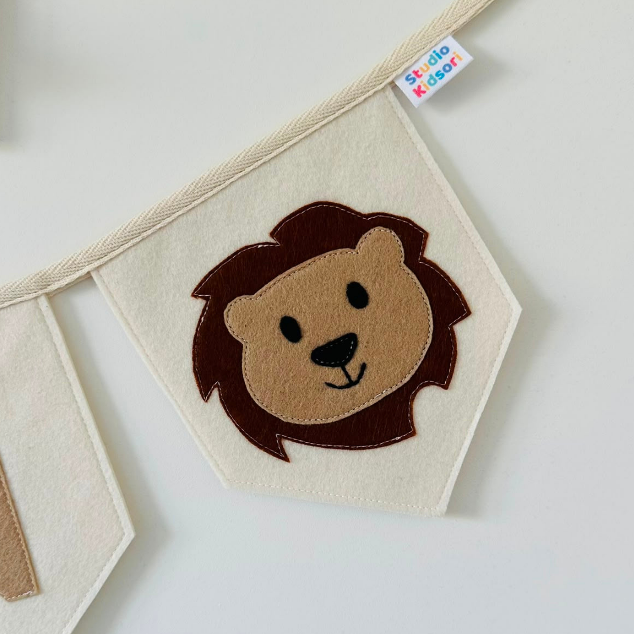Handgemaakte vilten vlag met een bruin beige leeuwenhoofd icoon op een champagne kleurige vlag, hangend aan een naturel katoenen lint. De leeuw heeft beige gezicht, zwarte ogen, neus en mond. de randen van de vlag zijn met een rechte afwerking afgewerkt.