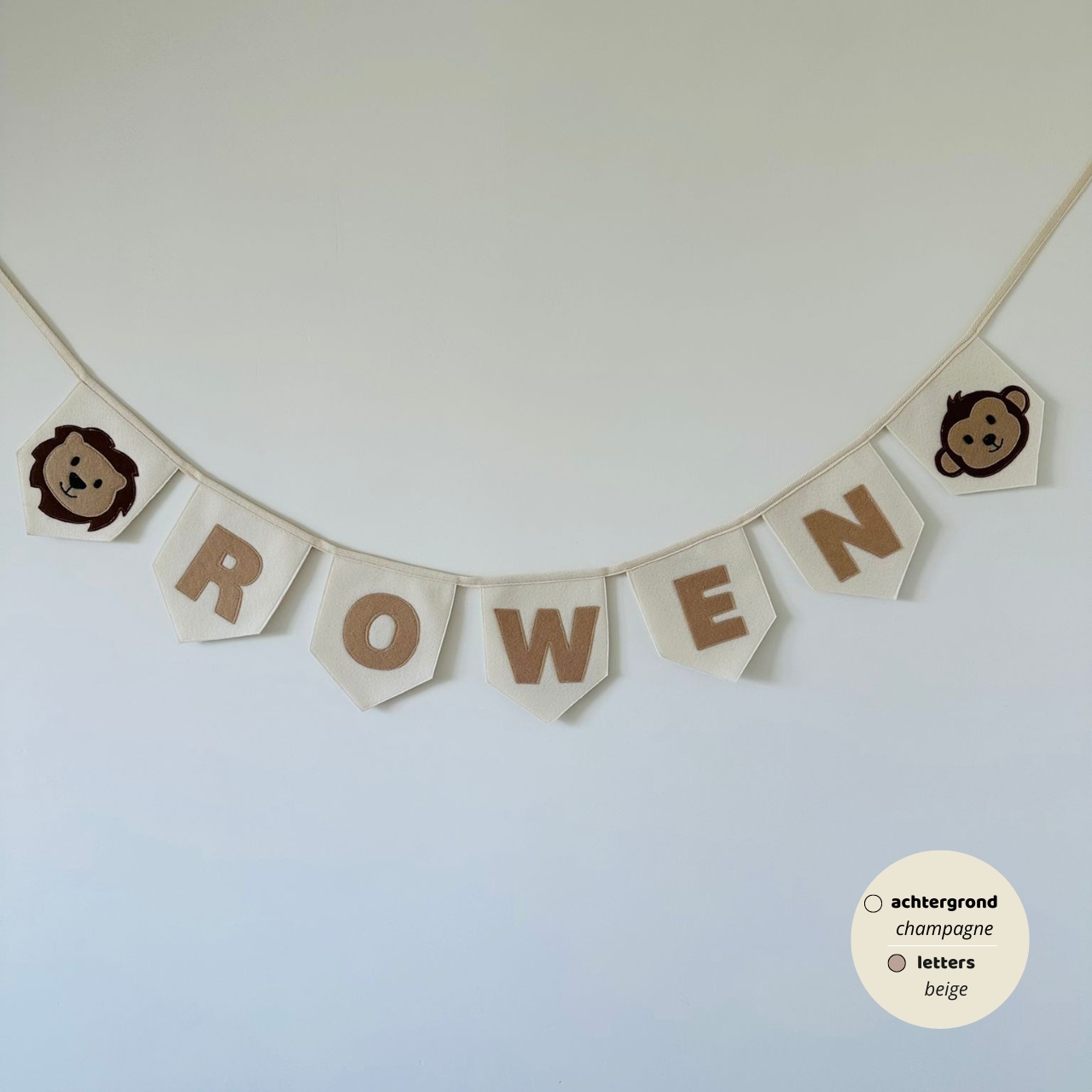 Gepersonaliseerde handgemaakte vilten naamvlaggenlijn met de naam "ROWEN", bestaande uit champagne kleurige vlaggen met beige vilten letters en bruin beige leeuwenhoofd op het eerste vlaggetje en apenhoofd  icoon op het laatste vlaggetje. De randen van de vlaggen zijn afgewerkt met een rechte afwerking. De vlaggenlijn hangt aan een naturel katoenen lint.