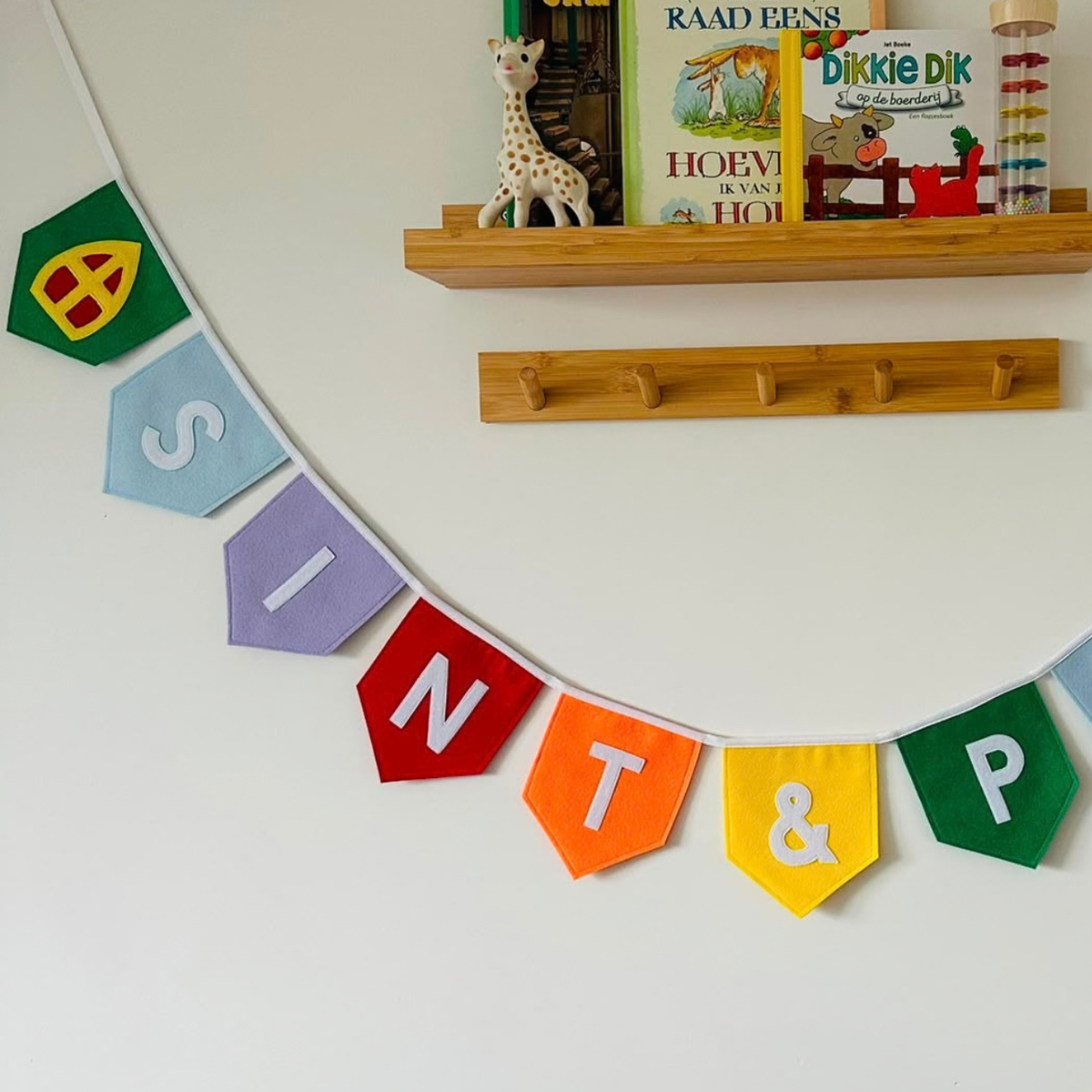 Sinterklaas vlaggenlijn van vilt met de letters Sint & P  in witte letters op gekleurde vlaggetjes, met aan de uiteinden een mijter en een Pietenmuts, hangend aan een muur boven een houten wandplank
