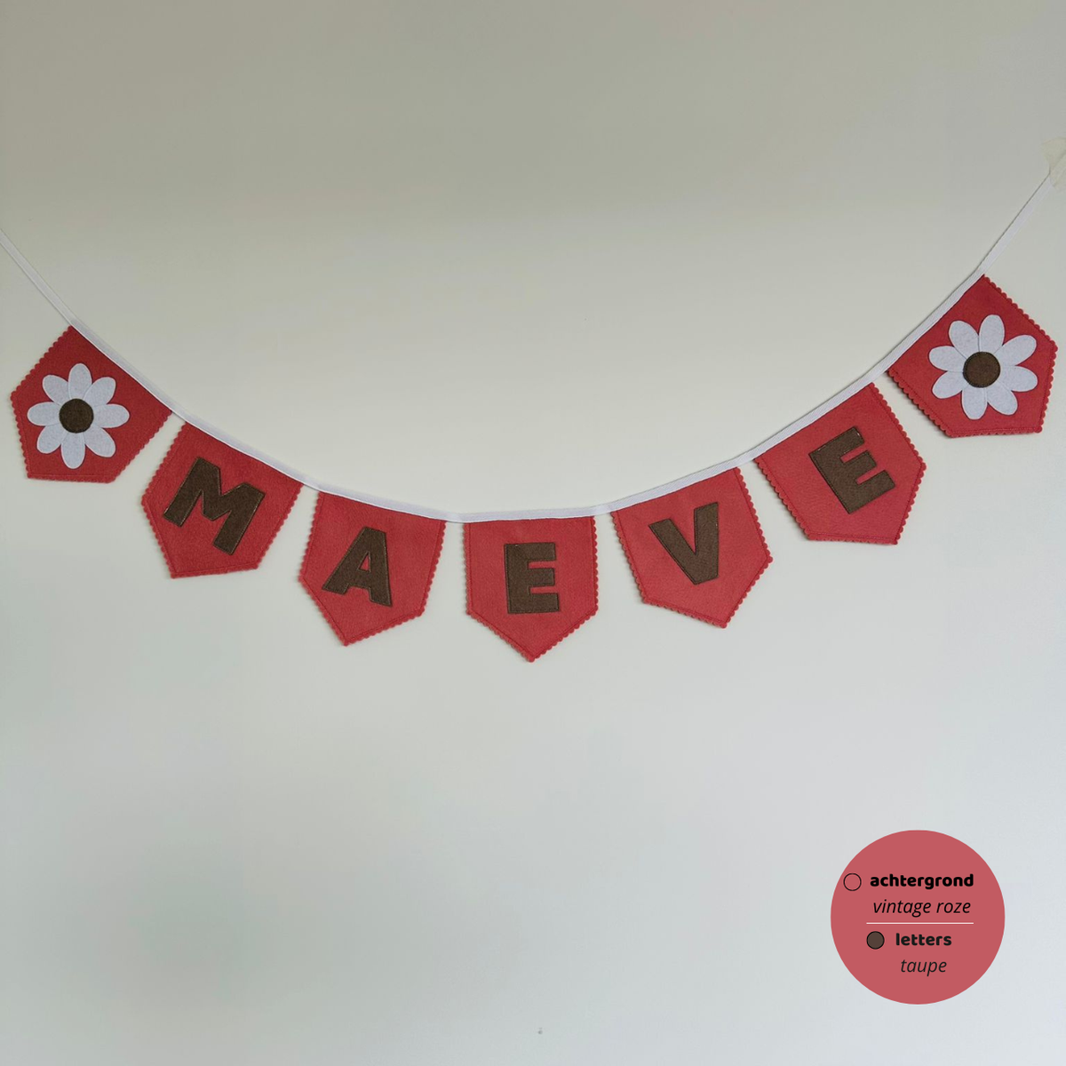 Gepersonaliseerde handgemaakte vilten naamvlaggenlijn met de naam "Maeve", bestaande uit vintage roze kleurige vlaggen met taupe vilten letters en wit met taupe bloem op het eerste vlaggetje en het laatste vlaggetje. De randen van de vlaggen zijn afgewerkt met een scallop afwerking. De vlaggenlijn hangt aan een wit katoenen lint.