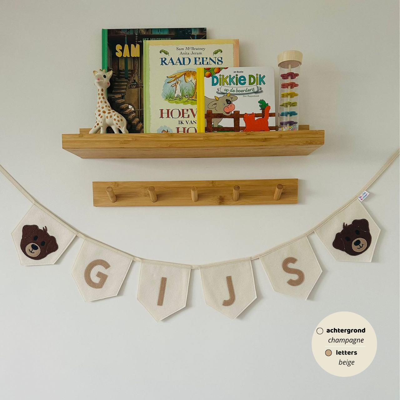 Gepersonaliseerde handgemaakte vilten naamvlaggenlijn met de naam "GIJS", bestaande uit champagne kleurige vlaggen met beige vilten letters en bruin hondenhoofd iconen met beige snoet op de eerste en laatste vlag. De randen van de vlaggen zijn afgewerkt met een rechte rand. De vlaggenlijn hangt aan een naturel katoenen lint in een kinderkamer, gefotografeerd onder een houten boekenplank met kinderboeken en een Sophie de Giraf speeltje, boven een houten kapstok.
