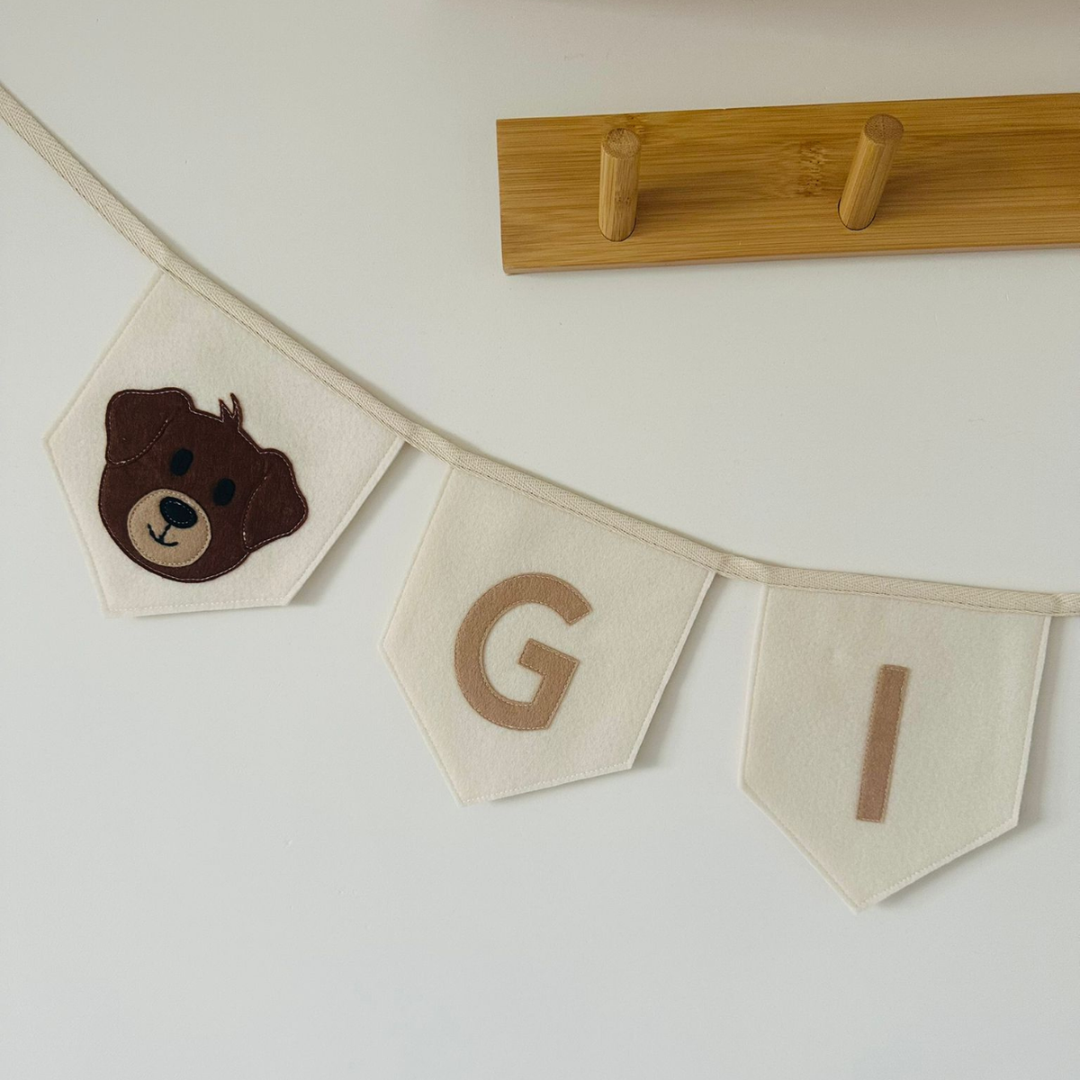 Gepersonaliseerde handgemaakte vilten naamvlaggenlijn met de eerste 2 letters van Gijs, bestaande uit champagne kleurige vlaggen met beige vilten letters en een bruin honden hoofd icoon met beige snoet op de eerste vlag. De randen van de vlaggen zijn afgewerkt met een rechte rand. De vlaggenlijn hangt aan een naturel katoenen lint, gefotografeerd tegen een lichte muur met een houten kapstok op de achtergrond.