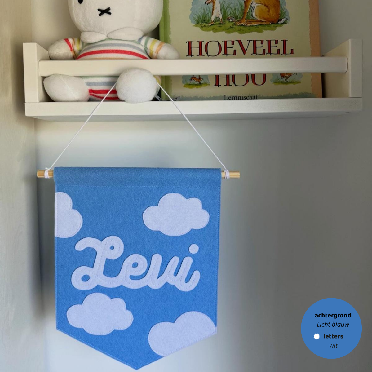 Gepersonaliseerde handgemaakte vilten banner met de naam "Levi", bestaande uit licht blauw kleurige banner met witte vilten letters wolken iconen. De randen van de banner is afgewerkt met een rechte rand. De banner hangt aan een wit katoenen koord aan een houten stok in een 