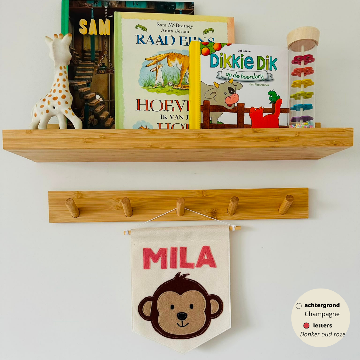 Gepersonaliseerde handgemaakte vilten banner met de naam "MILA", bestaande uit champagne kleurige banner met Donker oud roze vilten letters en aap icoon. De randen van de banner is afgewerkt met een rechte rand. De banner hangt aan een wit katoenen koord aan een houten stok in een kinderkamer.