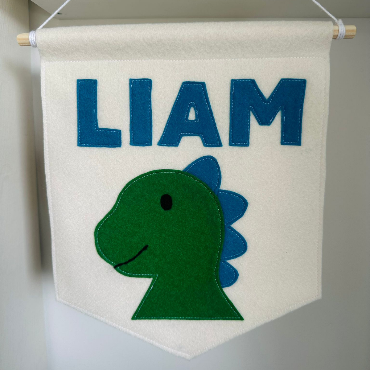 Gepersonaliseerde handgemaakte vilten banner met de naam "Liam", bestaande uit champagne kleurige banner met petroleum blauwe vilten letters en dino hoofd  icoon. De randen van de banner is afgewerkt met een rechte rand. De banner hangt aan een wit katoenen koord aan een houten stok in een kinderkamer.