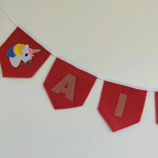 Close-up van een handgemaakte vintage roze vilten eenhoorn vlaggenlijn met naam, met een gedetailleerd eenhoorn-icoon met glitter hoorn en de letters "A" en "I" in oud roze vilt.