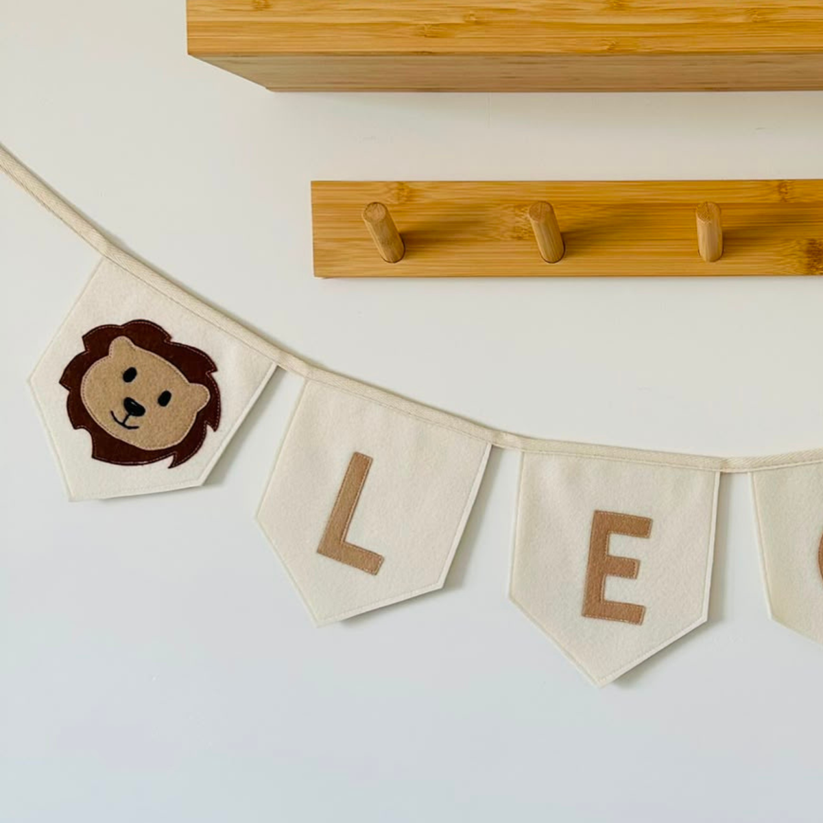 Close-up van een handgemaakte champagne vilten leeuw vlaggenlijn met naam, met een bruin leeuwenkop-icoon en de letters "L" en "E" in beige vilt.