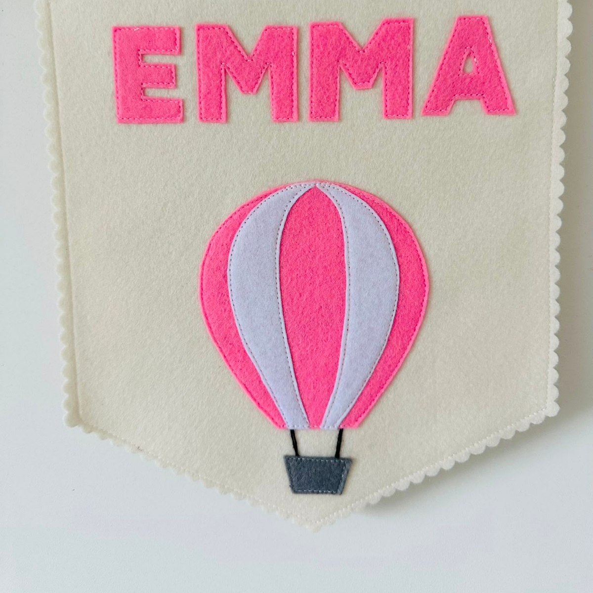 Gepersonaliseerde handgemaakte vilten banner met de naam "Emma", bestaande uit champagne kleurige banner met pastel roze vilten letters en luchtballon icoon. De randen van de banner is afgewerkt met een scallop rand. De banner hangt aan een wit katoenen koord aan een houten stok in een kinderkamer.
