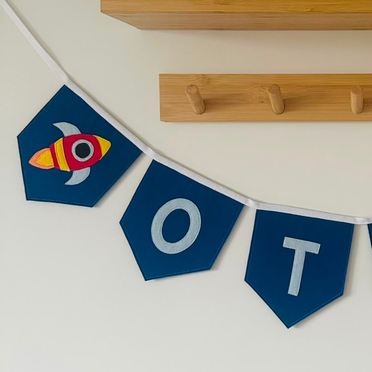 Close-up van een handgemaakte donkerblauwe vilten raket vlaggenlijn met naam, met een kleurrijk raket-icoon en de letters "O" en "T" in lichtblauw vilt.