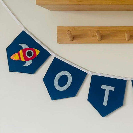 Close-up van een handgemaakte donkerblauwe vilten raket vlaggenlijn met naam, met een kleurrijk raket-icoon en de letters "O" en "T" in lichtblauw vilt.