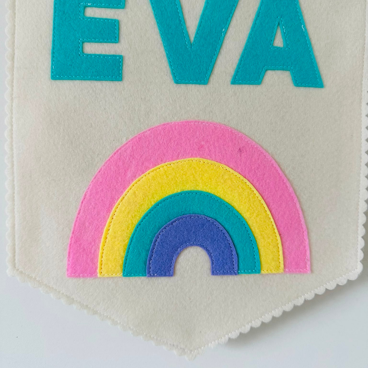 Gepersonaliseerde handgemaakte vilten banner met de naam "EVA", bestaande uit champagne kleurige banner met pastel groene vilten letters en regenboog icoon. De randen van de banner is afgewerkt met een scallop rand. De banner hangt aan een wit katoenen koord aan een houten stok in een kinderkamer.