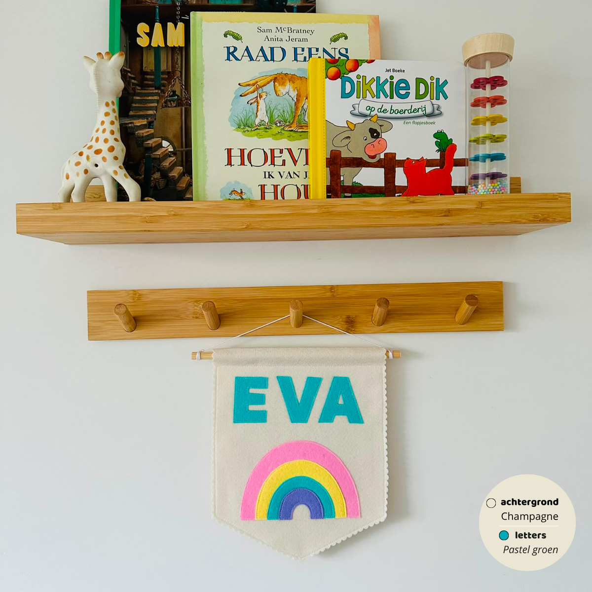 Gepersonaliseerde handgemaakte vilten banner met de naam "EVA", bestaande uit champagne kleurige banner met pastel groene vilten letters en regenboog icoon. De randen van de banner is afgewerkt met een scallop rand. De banner hangt aan een wit katoenen koord aan een houten stok in een kinderkamer.