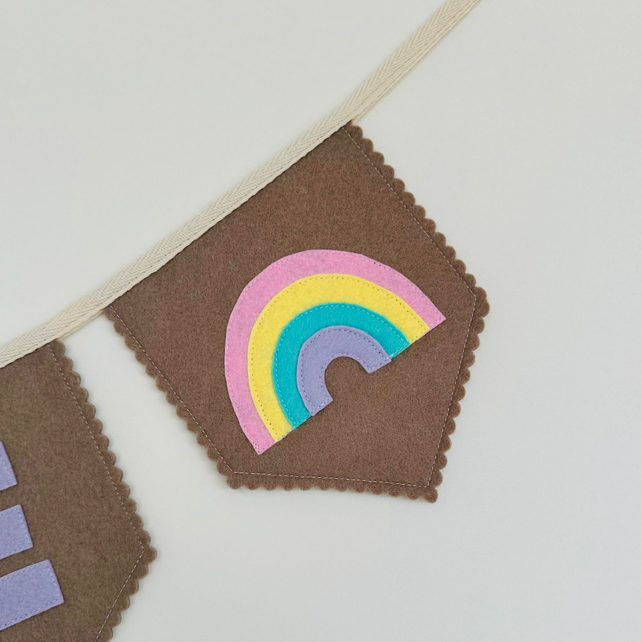 Handgemaakte vilten vlag met een wregenboog icoon op, hangend aan een witte katoenen lint. De randen van de vlag zijn met een scalp afwerking afgewerkt.