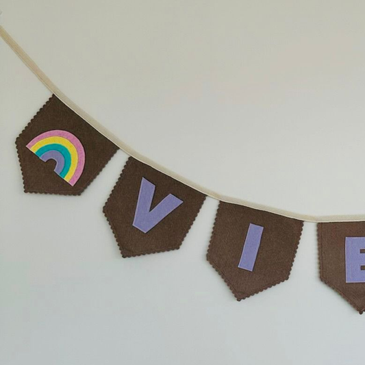 Close-up van een handgemaakte taupe vilten regenboog vlaggenlijn met naam, met een kleurrijke regenboog-icoon en de letters "V" en "I" in lila vilt.