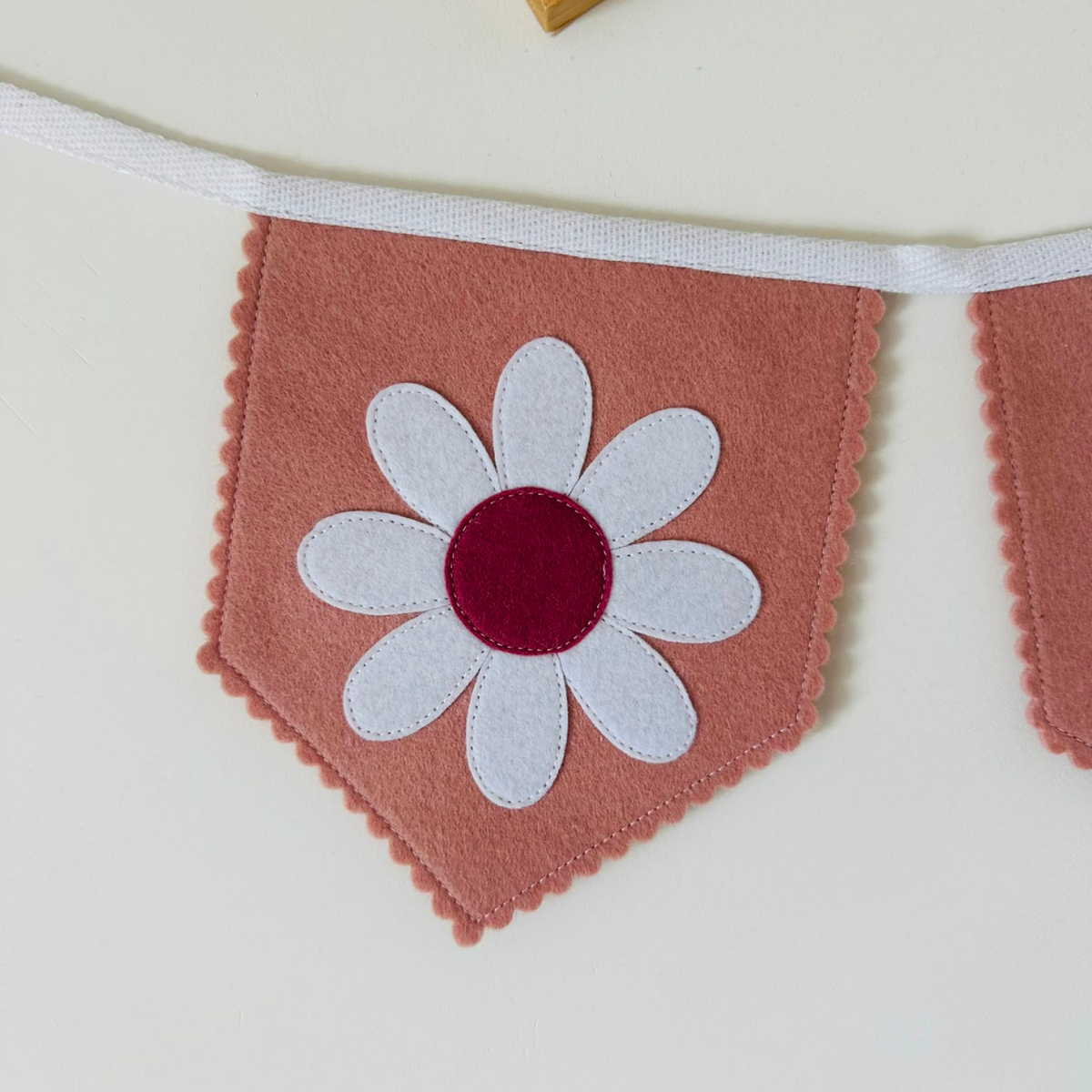 Handgemaakte vilten vlag met een wit en donker oud roze bloem icoon op een oud roze kleurige vlag, hangend aan een witte katoenen lint. De randen van de vlag zijn met een scalp afwerking afgewerkt.