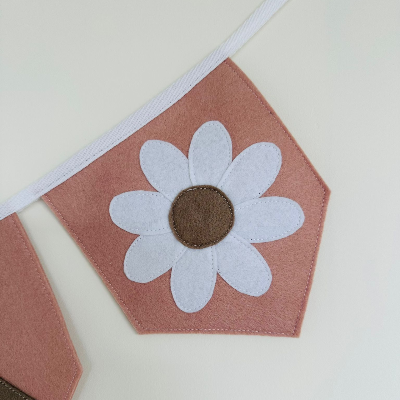 Handgemaakte vilten vlag met een wit en taupe bloem icoon op een oud roze kleurige vlag, hangend aan een witte katoenen lint. De randen van de vlag zijn met een rechte afwerking afgewerkt.