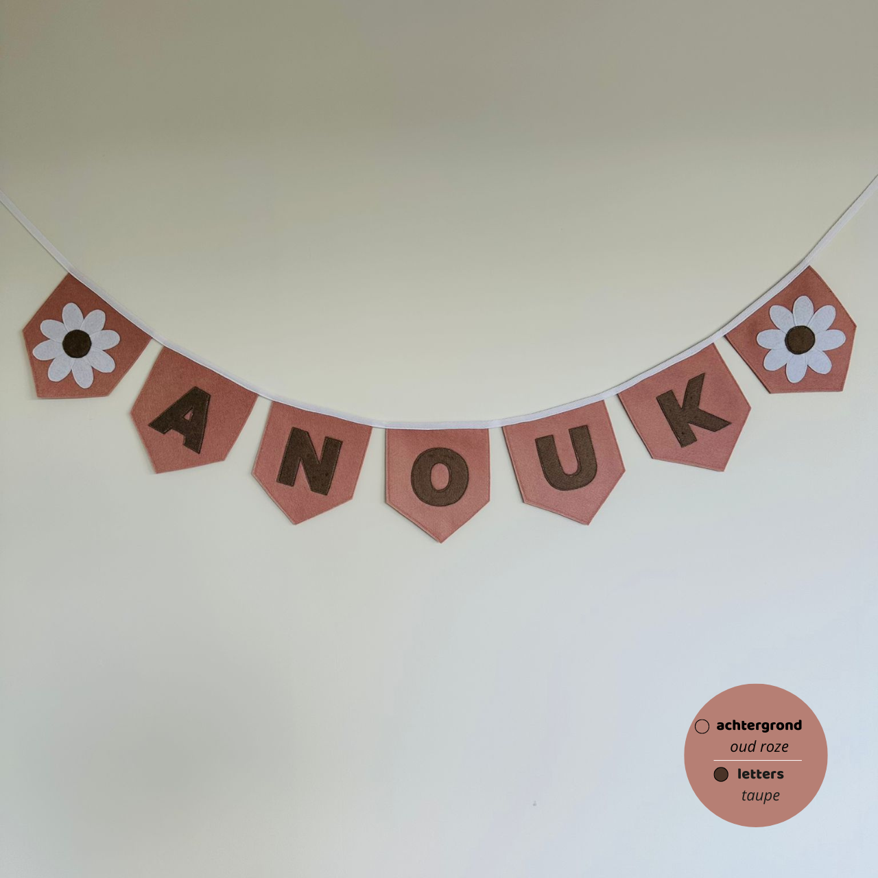 Gepersonaliseerde handgemaakte vilten naamvlaggenlijn met de naam "ANOUK", bestaande uit oud roze kleurige vlaggen met taupe vilten letters en wit met taupe bloemop het eerste vlaggetje en het laatste vlaggetje. De randen van de vlaggen zijn afgewerkt met een rechte afwerking. De vlaggenlijn hangt aan een wit katoenen lint.
